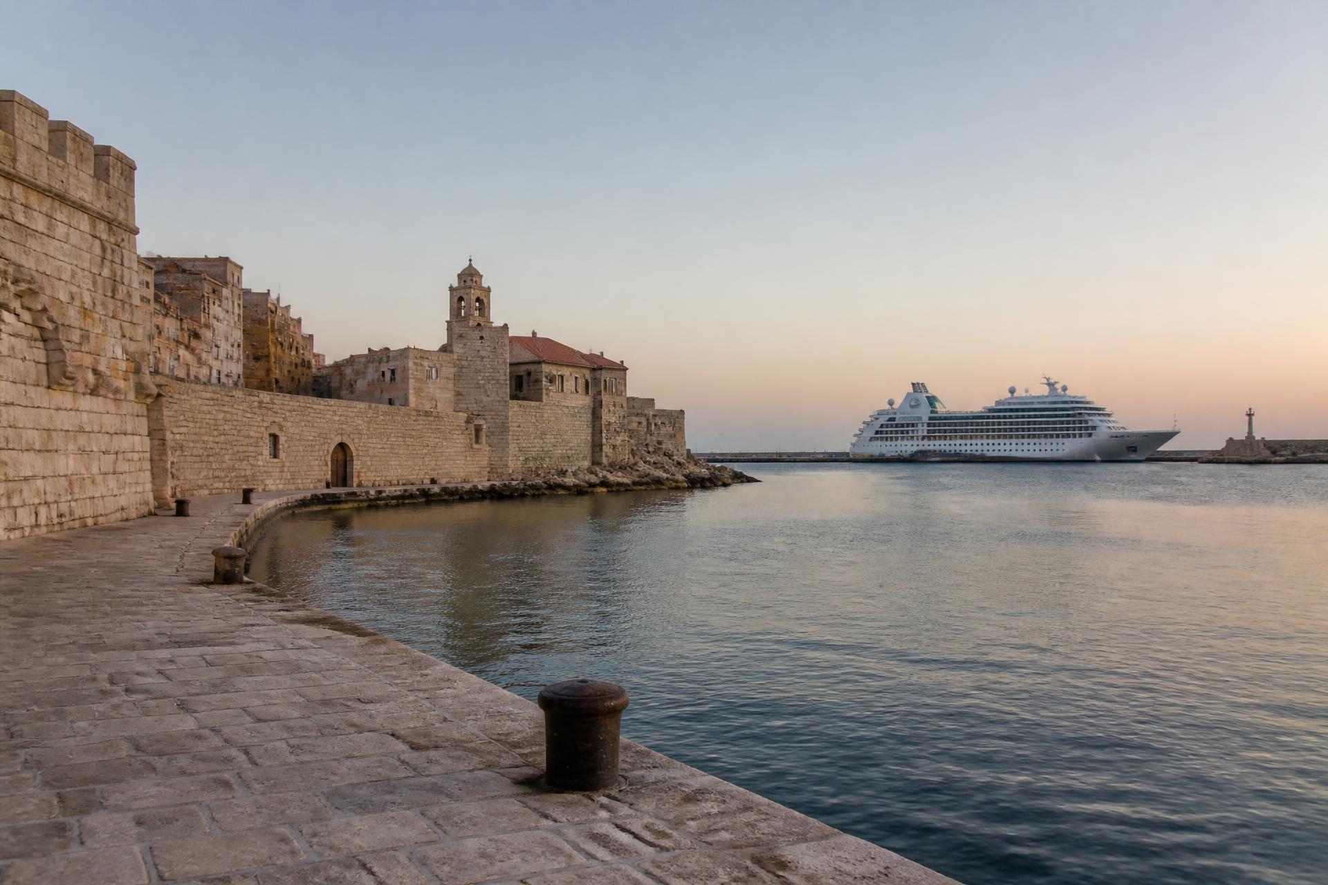 A stone harbour in soft evening light with a cruise ship berthed beyond the wall